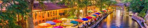 Colorful umbrellas line a riverside walkway, illuminated by warm lights, creating a vibrant evening atmosphere.