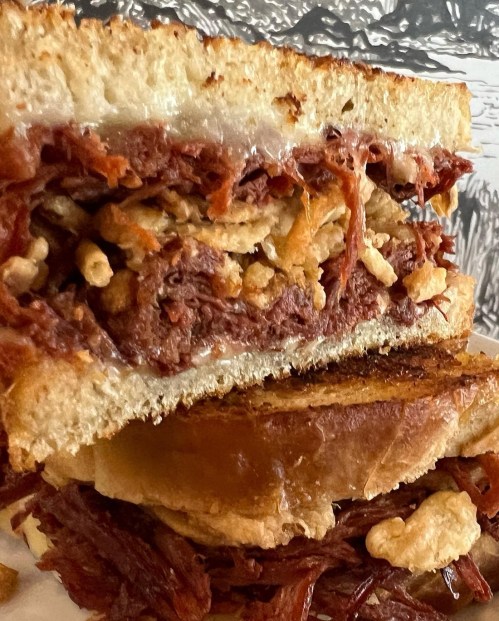 A close-up of a stacked sandwich filled with shredded meat, crispy onions, and melted cheese on toasted bread.