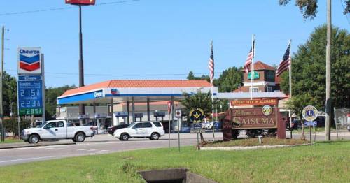Welcome sign for Satsuma, Alabama, with a Chevron gas station and flags in the background.
