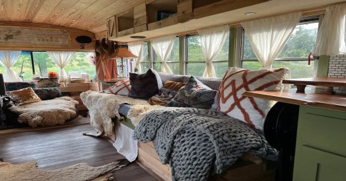 Cozy interior of a bus converted into a living space, featuring plush pillows, blankets, and large windows with natural light.