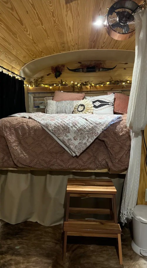 Cozy interior of a tiny home with a bed, decorative pillows, fairy lights, and a wooden stool. Warm, inviting atmosphere.