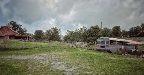A rustic scene featuring a red barn, a gray bus, and a grassy field under a cloudy sky.