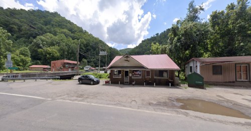A small wooden building with a red roof, surrounded by greenery and mountains, with a car parked in front.