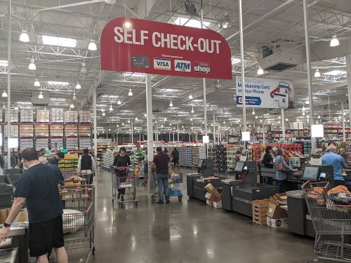 A spacious grocery store interior with self-checkout stations and shoppers with carts. Brightly lit aisles in the background.