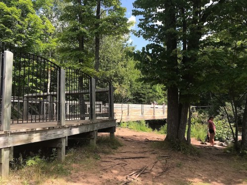 A wooden bridge surrounded by trees, with a person standing near the water's edge on a sunny day.