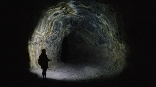 A person stands in a dark cave, illuminated by a flashlight, revealing textured rock walls and a shadowy entrance.