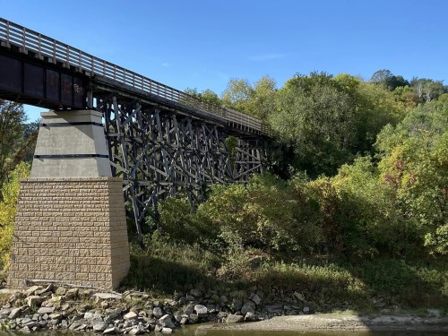 A wooden bridge supported by stone and wooden pillars, surrounded by lush greenery and a clear blue sky.