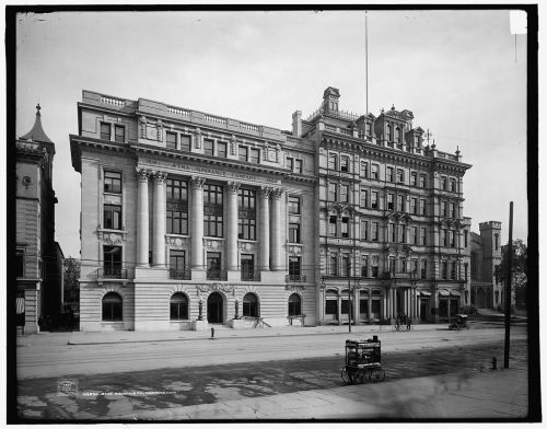 Historic black-and-white photo of a grand building with columns, featuring intricate architectural details and a street view.