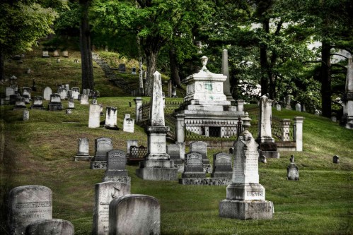 A serene cemetery with various gravestones and monuments set on a grassy hillside, surrounded by trees.
