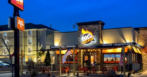 A brightly lit restaurant named "Big Chicken" with outdoor seating, flags, and a welcoming atmosphere at dusk.