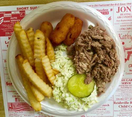 A plate of pulled pork, coleslaw, crinkle-cut fries, and hush puppies on a printed paper placemat.