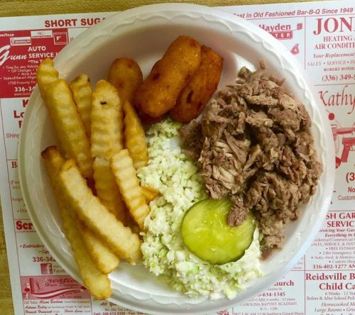 A plate of pulled pork, coleslaw, crinkle-cut fries, and hush puppies on a printed paper placemat.