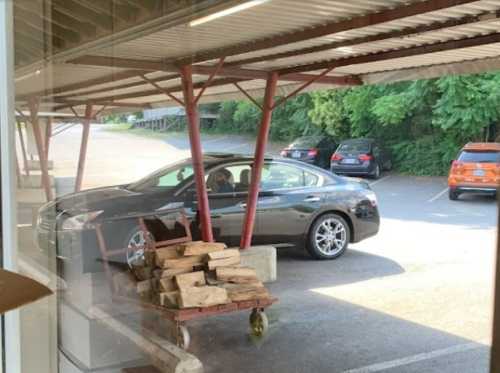 A gray car parked under a shelter, with a stack of firewood in the foreground and other cars in the background.