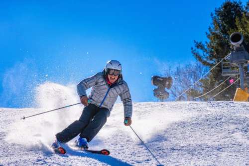 A skier in a gray jacket carves down a snowy slope, kicking up powder against a clear blue sky.