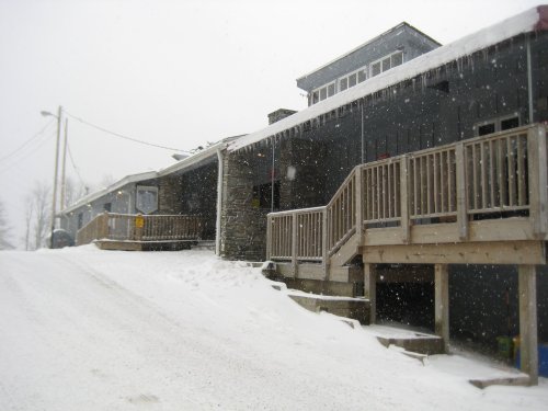 Snowy scene of a row of buildings with wooden decks, surrounded by a winter landscape and falling snowflakes.