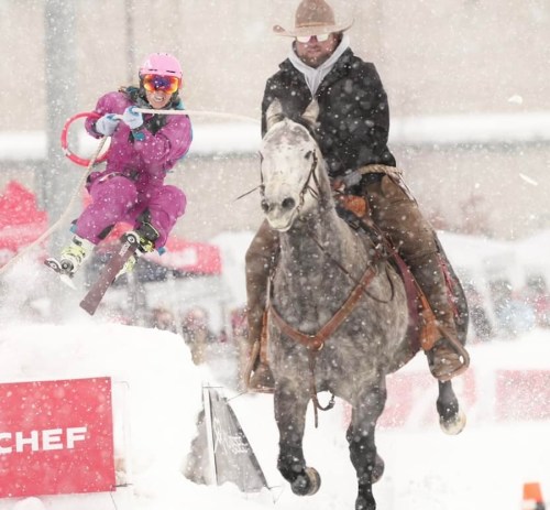 A skier in colorful gear jumps over a snow ramp while being pulled by a horse, with snow falling around them.