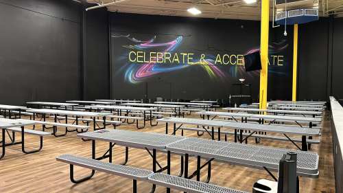 A spacious indoor area with rows of metal picnic tables and a colorful mural that says "CELEBRATE & ACCELERATE."