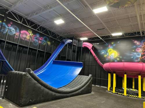 Indoor play area featuring a blue slide and a pink slide, with colorful wall art in the background.