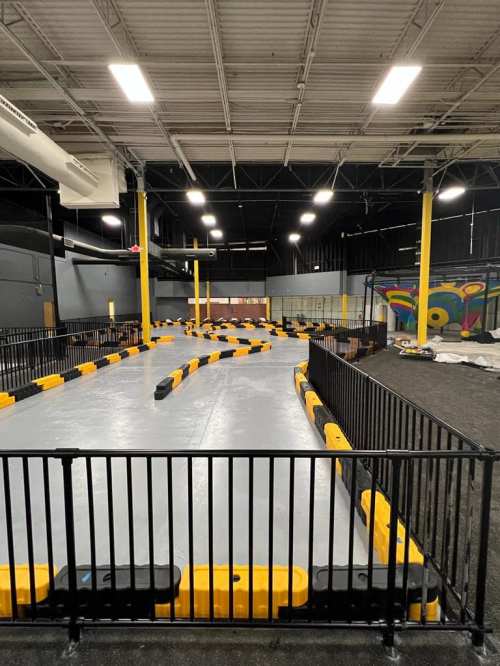Indoor go-kart track with a winding path, surrounded by black fencing and yellow barriers, under bright overhead lights.