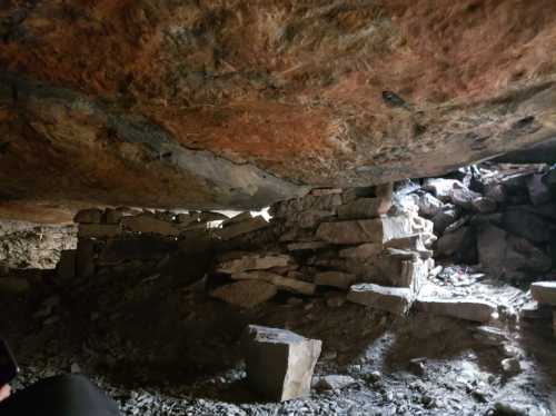 A dimly lit cave interior with rough stone walls and a low ceiling, revealing a small opening to the outside.