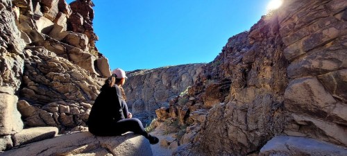 A person sits on a rock ledge, gazing at a canyon with steep walls under a clear blue sky.