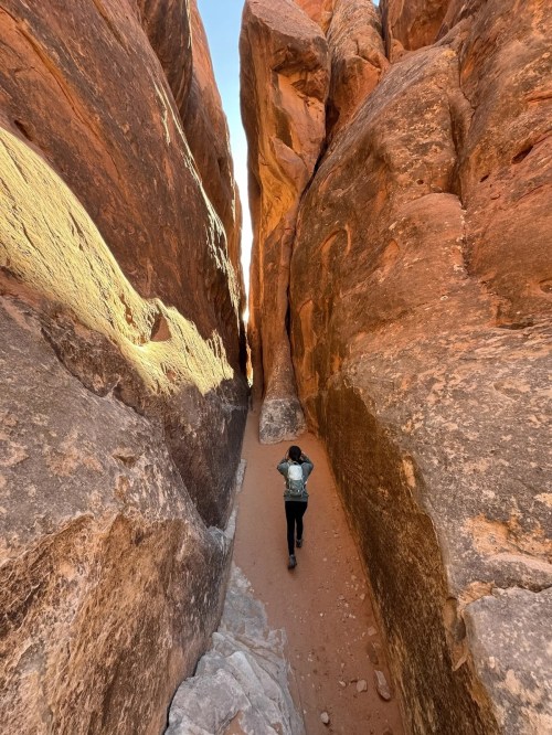 A person walks through a narrow canyon surrounded by towering red rock formations.