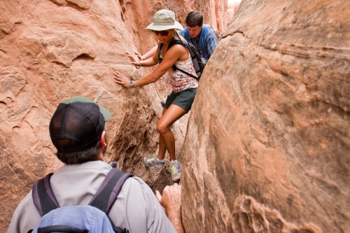 Three hikers navigate a narrow canyon, with one climbing while the others assist from below.