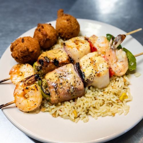 A plate of grilled shrimp and seafood skewers served over rice, accompanied by two fried balls.