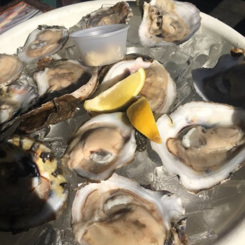 A platter of fresh oysters on ice, accompanied by lemon wedges and a small cup of sauce.