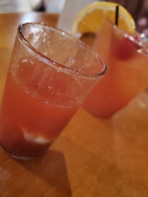 Two glasses of colorful cocktails with ice, garnished with a slice of orange and a cherry, on a wooden table.