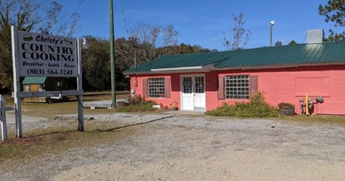 A pink building with a green metal roof, labeled "Christy's Country Cooking," surrounded by gravel and trees.