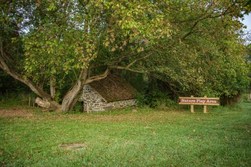 A small stone building under a tree, next to a sign that reads "Nature Play Area," surrounded by grass and foliage.