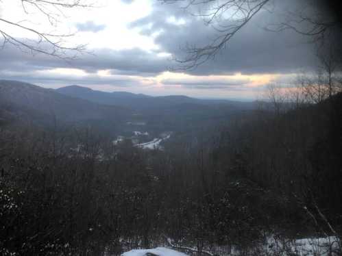 A scenic view of mountains and valleys under a cloudy sky, with hints of sunset and snow on the ground.