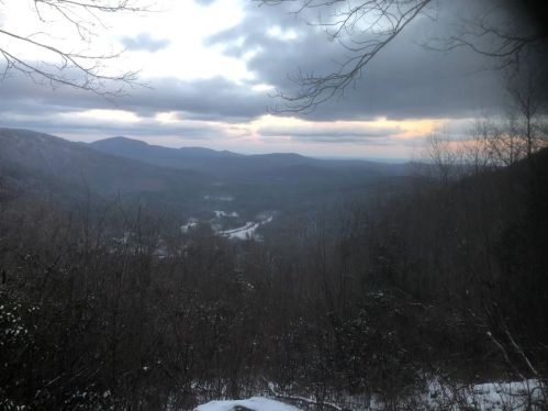 A scenic view of mountains and valleys under a cloudy sky, with hints of sunset and snow on the ground.