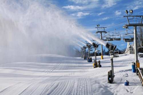 Snowmaking machines create artificial snow on a ski slope, with empty chairlifts in the background under a clear blue sky.