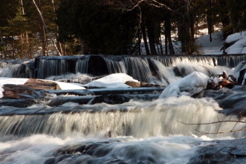 A serene waterfall cascades over rocks, surrounded by snow and trees in a tranquil forest setting.