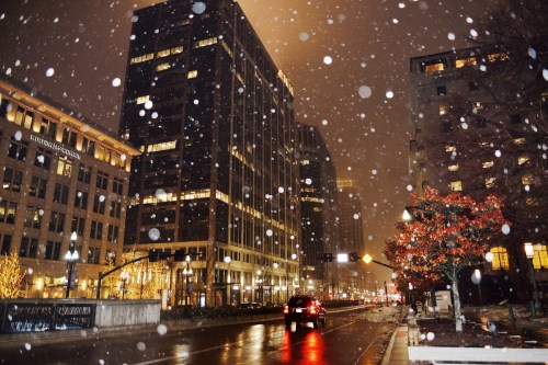 A snowy city street at night, with tall buildings and festive lights, creating a cozy winter atmosphere.