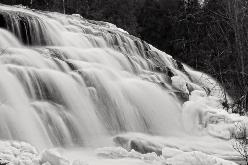 A cascading waterfall with smooth, flowing water and frothy white foam, surrounded by trees in a black and white setting.