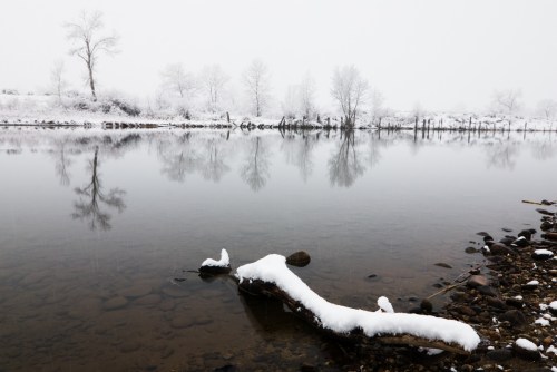 A snowy landscape with a calm river reflecting trees and a snow-covered log on the shore.
