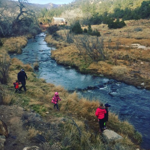 A family walks along a stream in a natural setting, with trees and mountains in the background.