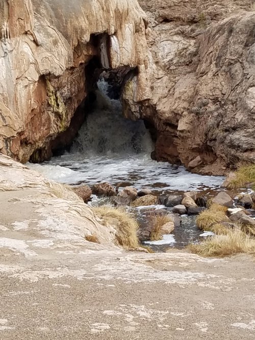A rocky landscape featuring a natural arch with water flowing through it, surrounded by boulders and grass.