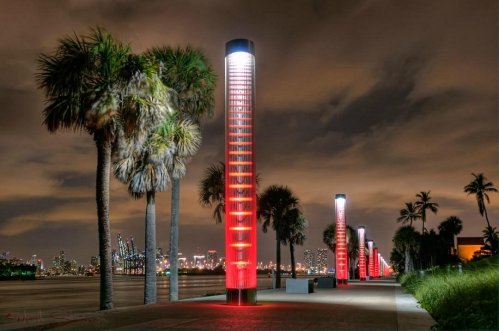 Night view of illuminated red columns along a waterfront, with palm trees and a city skyline in the background.