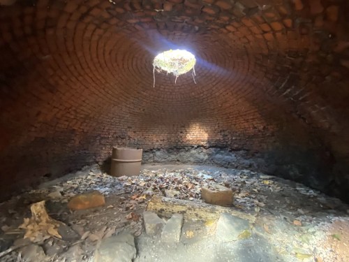 Interior of a brick structure with a circular opening at the top, surrounded by leaves and stones on the ground.