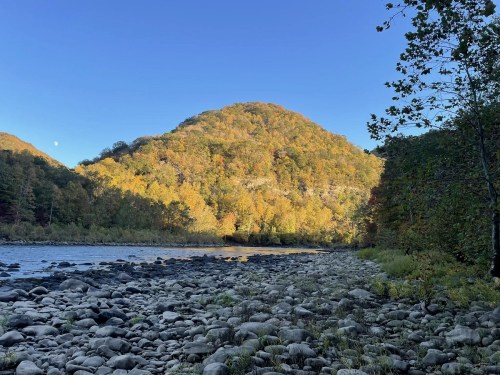 A serene riverbank with smooth stones, framed by a colorful hillside and clear blue sky.