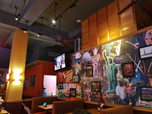 Interior of a restaurant featuring a colorful wall collage with New York landmarks and a TV in the background.