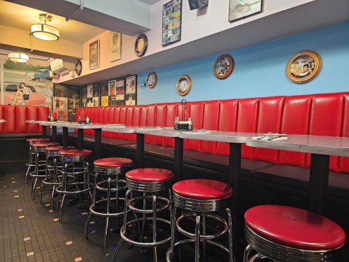 A retro diner interior featuring red booths, chrome stools, and vintage decor on the walls.