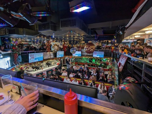 A bustling bar filled with people, decorated with lights and festive decor, viewed from an upper level.