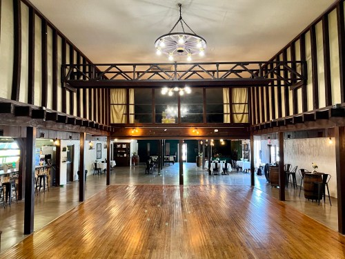 A spacious interior of a venue with wooden beams, a balcony, and a polished wooden floor, featuring tables and chairs.