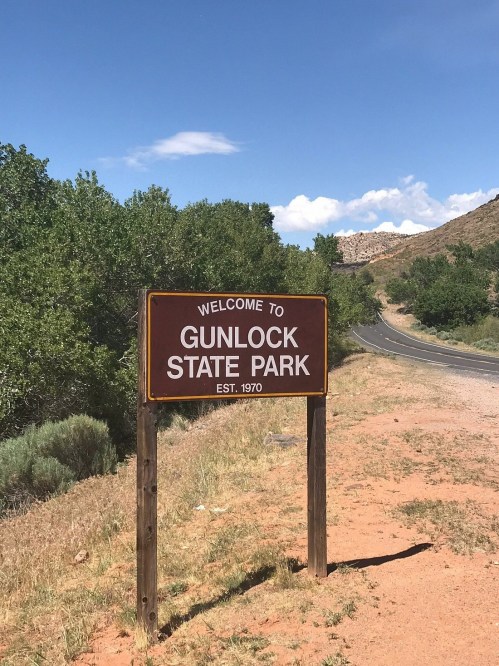 Sign welcoming visitors to Gunlock State Park, established in 1970, with a road and trees in the background.
