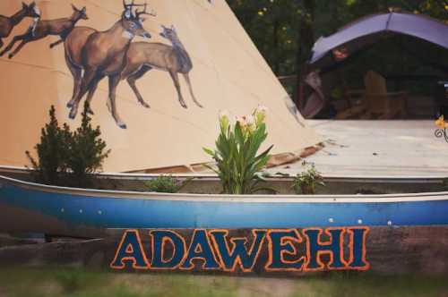 A wooden sign reading "ADAWEHI" in front of a canoe, with a teepee and deer mural in the background.